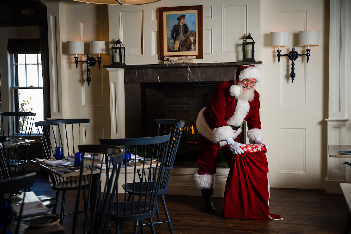 Santa Claus standing by a fireplace, reaching into a red gift sack in a dining room.