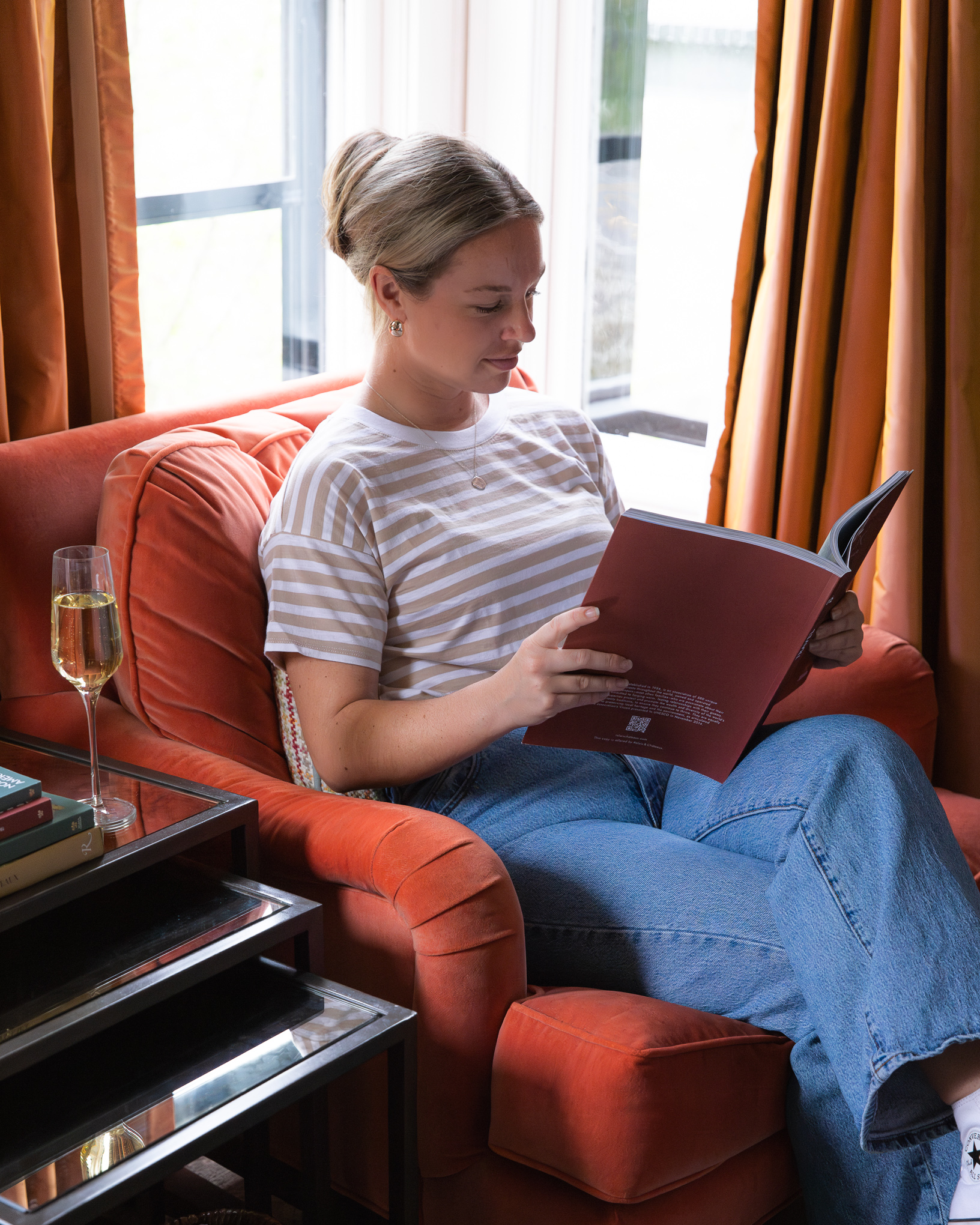 A woman sits in an orange armchair, reading a book, with a glass of champagne on a side table.