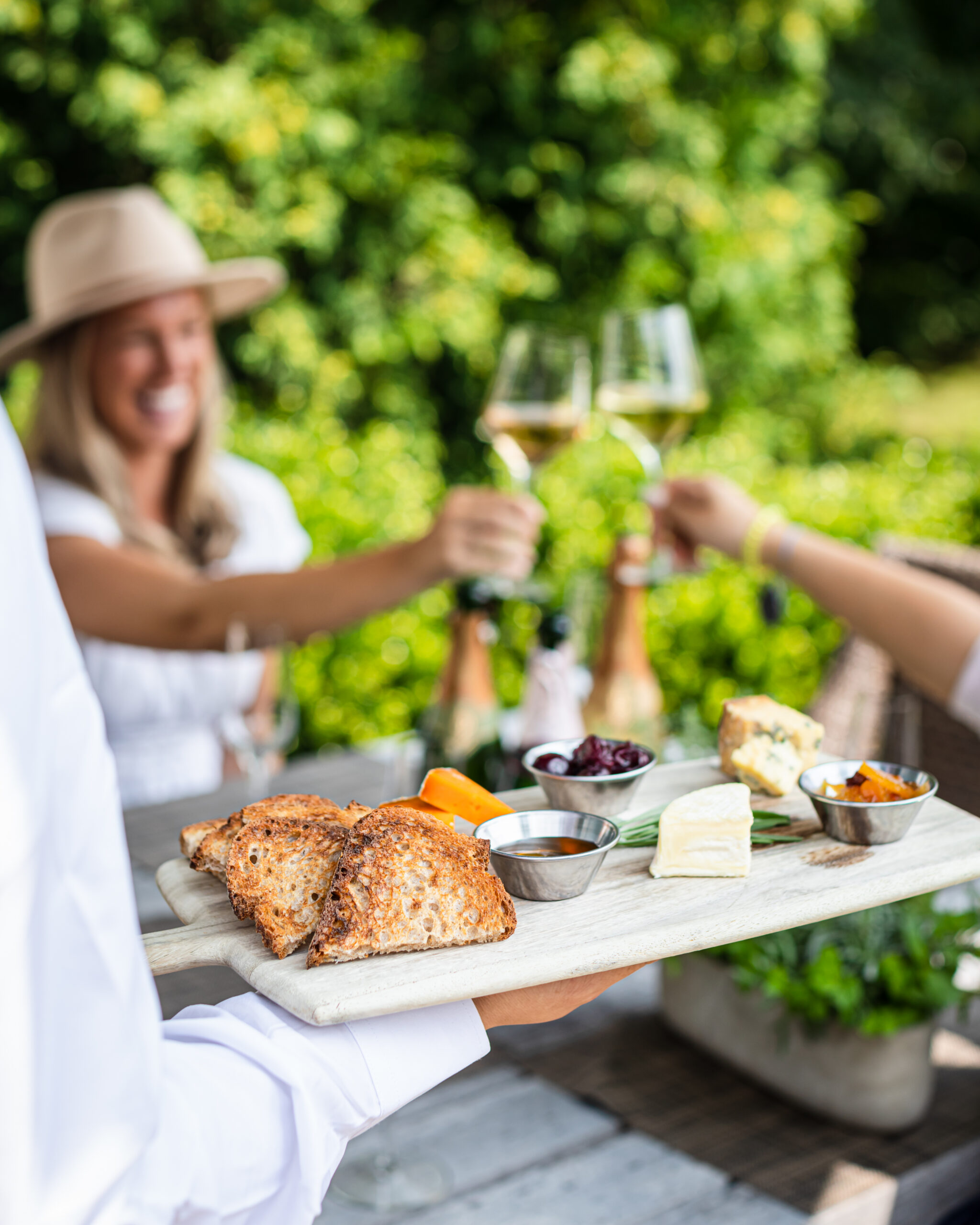 A person serves a cheese and bread platter as two people toast with wine outdoors, surrounded by greenery.