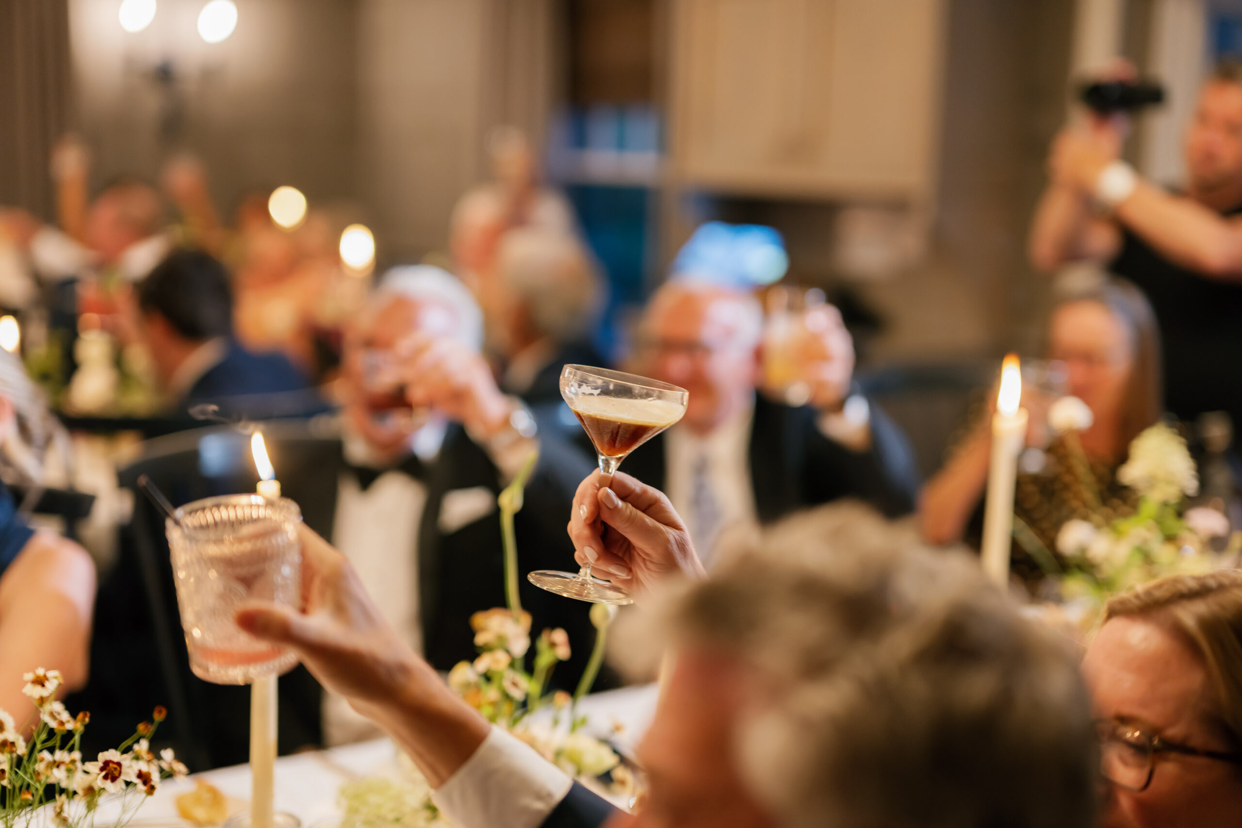 People dressed formally raise their glasses for a toast at a candlelit dinner event with floral table decorations.