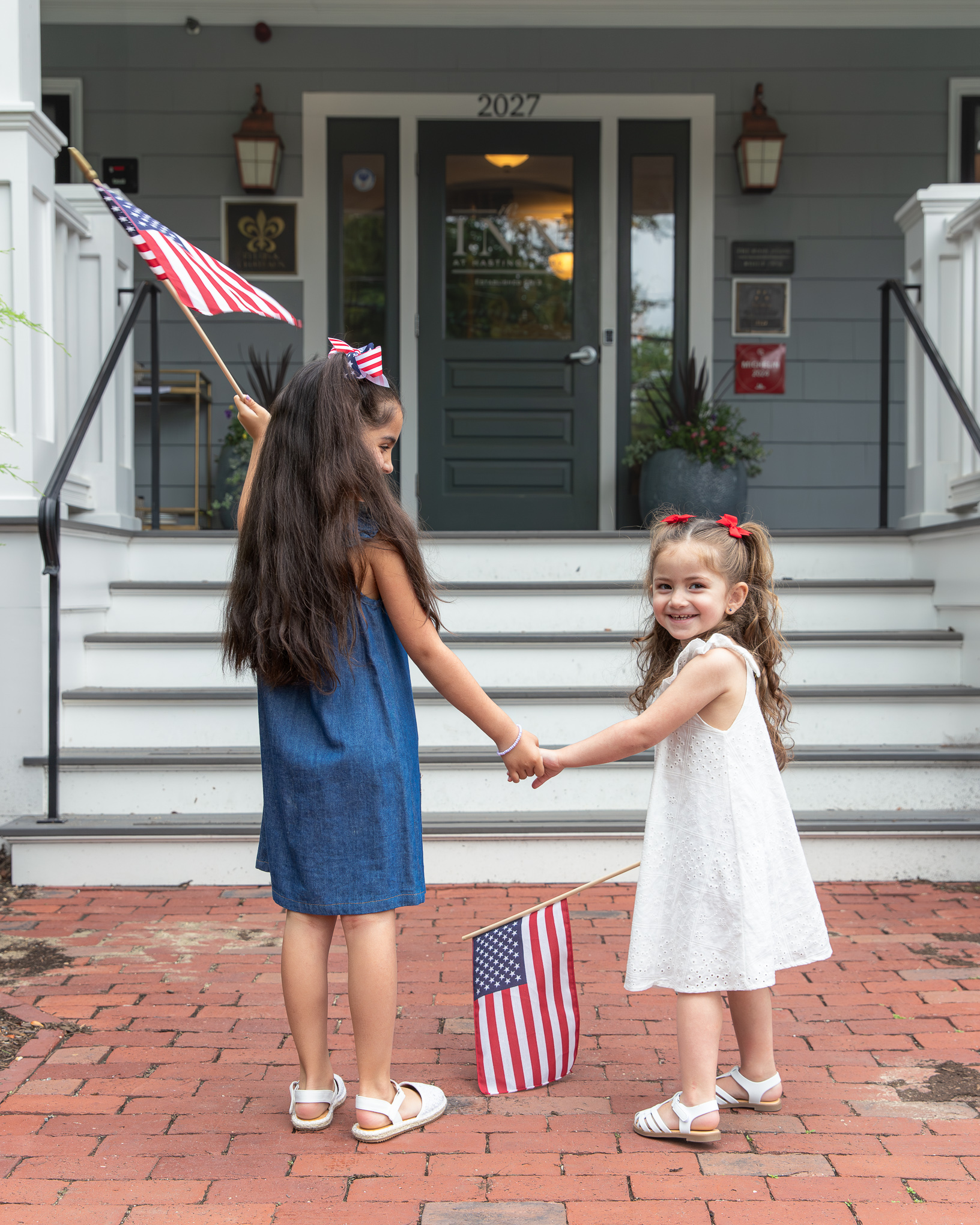 Two girls hold hands and American flags on brick pavement in front of a house.
