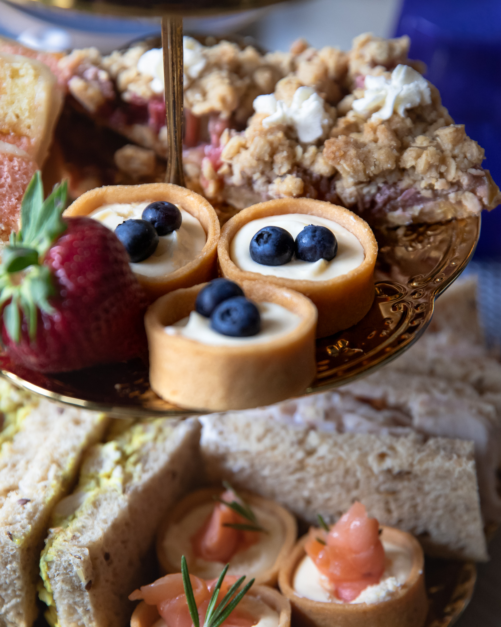 A gold tray with tarts topped with blueberries, a strawberry, crumble dessert, and assorted sandwiches arranged for afternoon tea.
