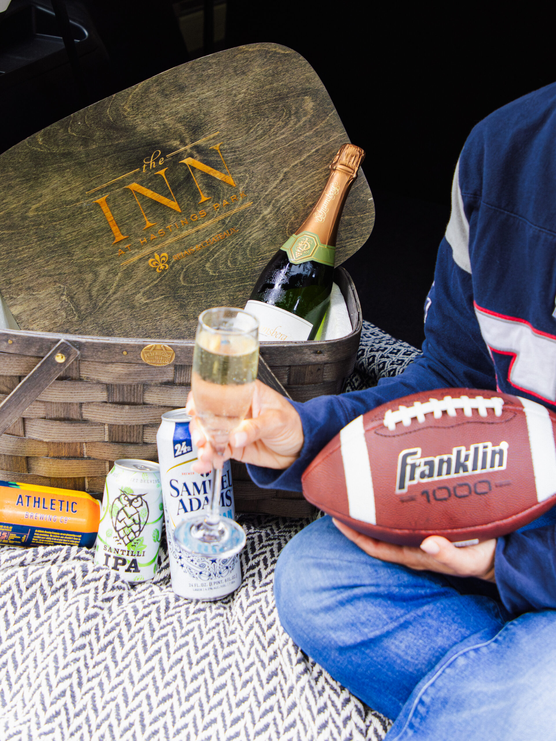 Person holding a football and champagne, seated on a picnic blanket with beer cans, a champagne bottle, and a basket labeled The Inn..