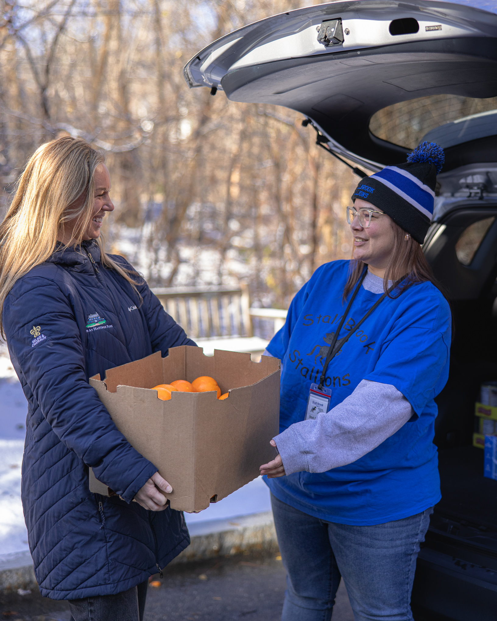 Two women smiling as one hands a box of oranges to the other by an open car trunk on a snowy day.