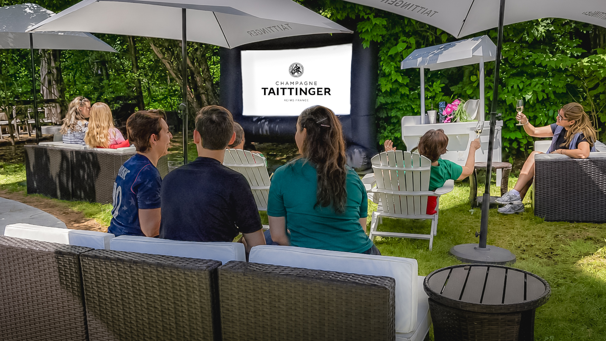 People watch an outdoor screen under umbrellas, surrounded by greenery. The screen displays Champagne Taittinger.