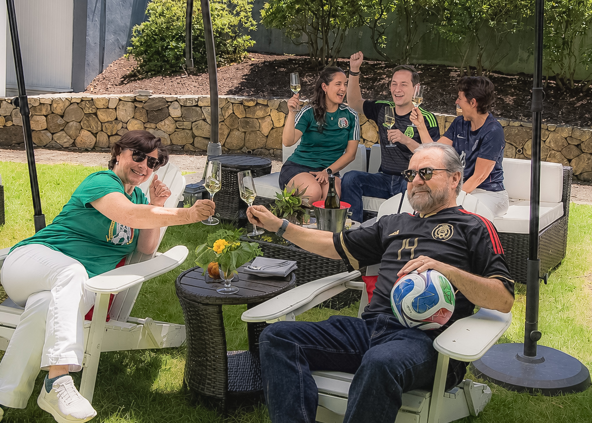 Two people in the foreground toast with drinks while three others chat and laugh behind them on a sunny lawn; a soccer ball rests on one persons lap.