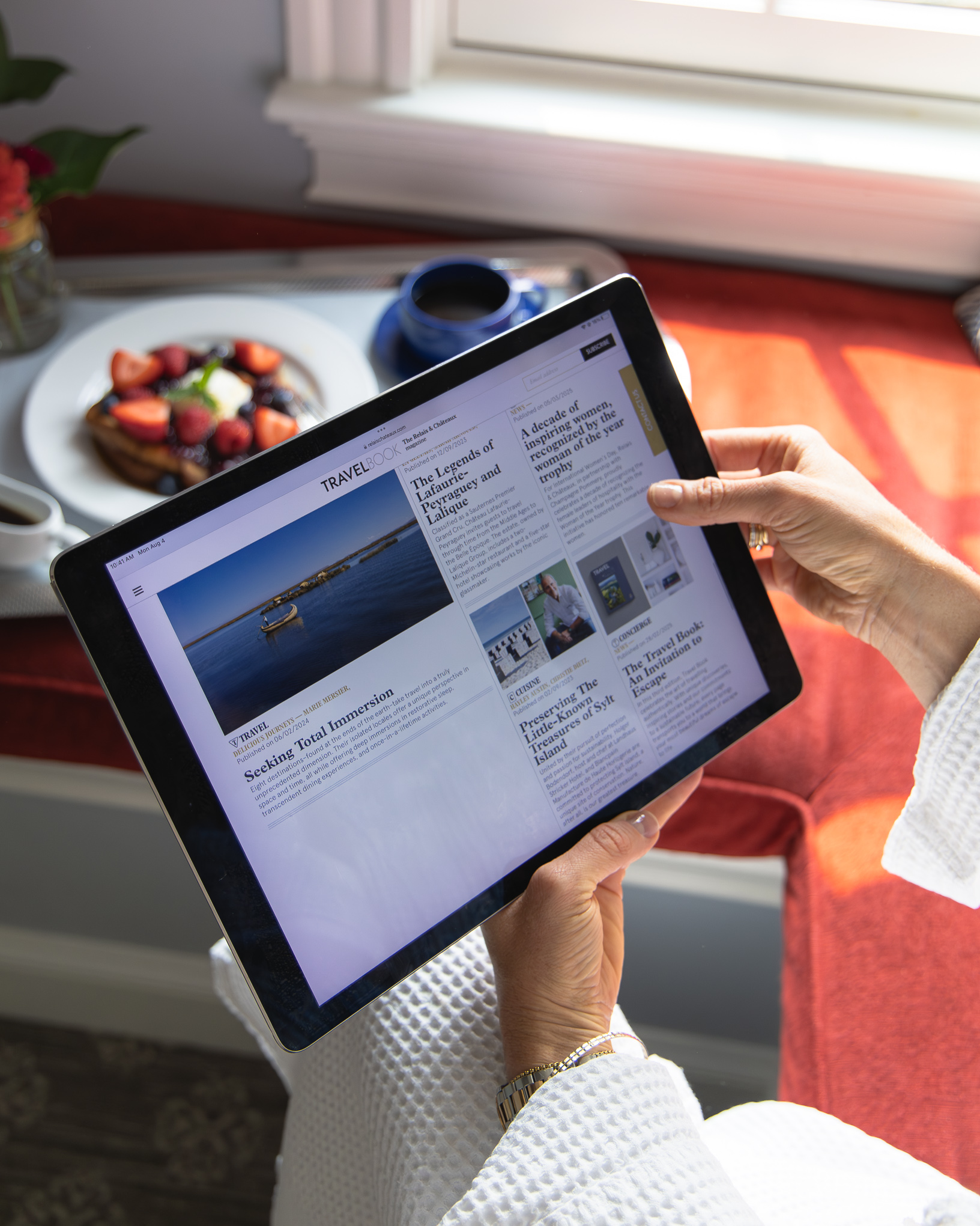 Person in a white robe holding a tablet displaying a travel news site, seated by a window with sunlight, coffee, and a plate of fruit nearby.