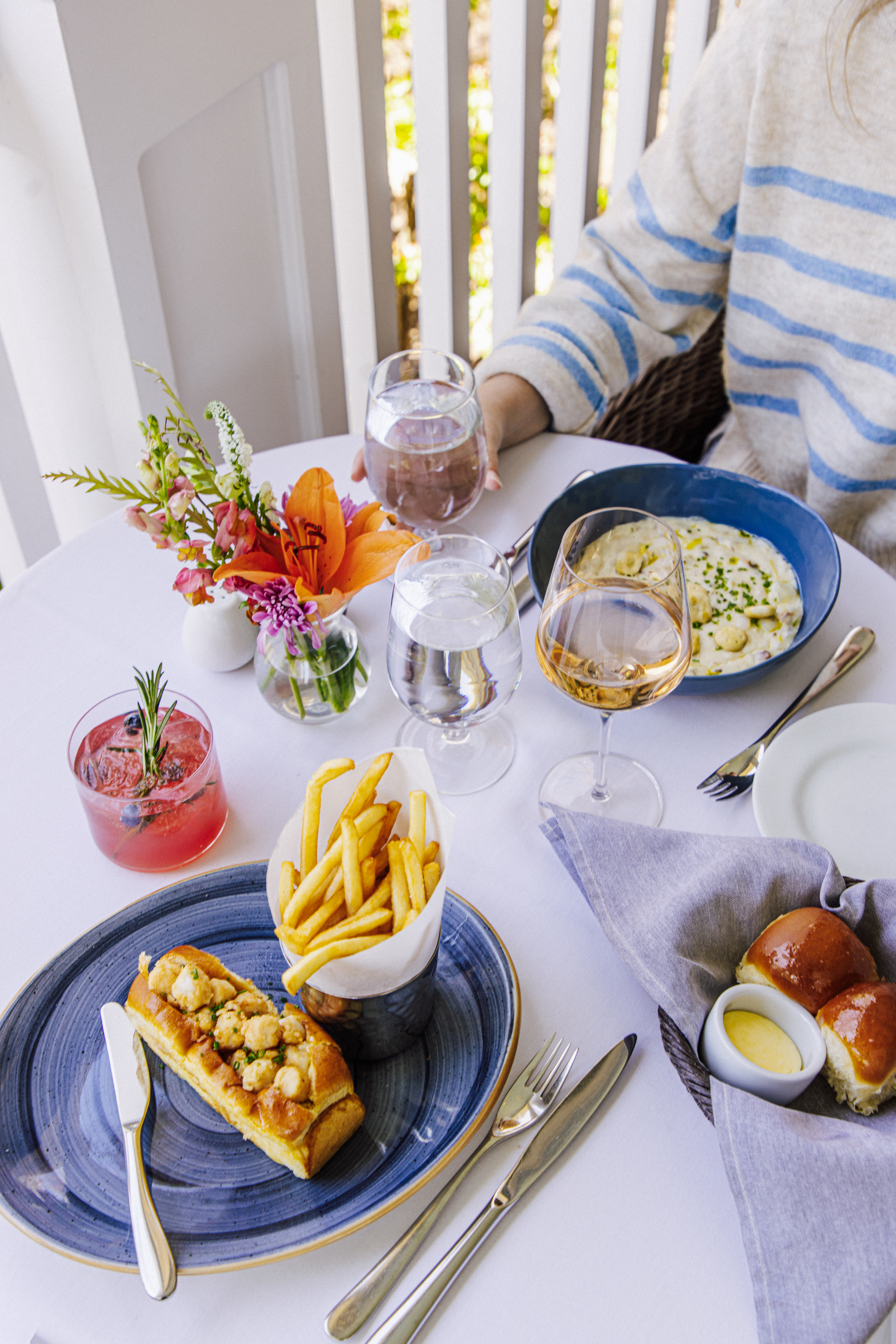 Table set with florals in a porch table with chowder, scallop roll with fries, waters, cocktail, rose wine and dinner rolls